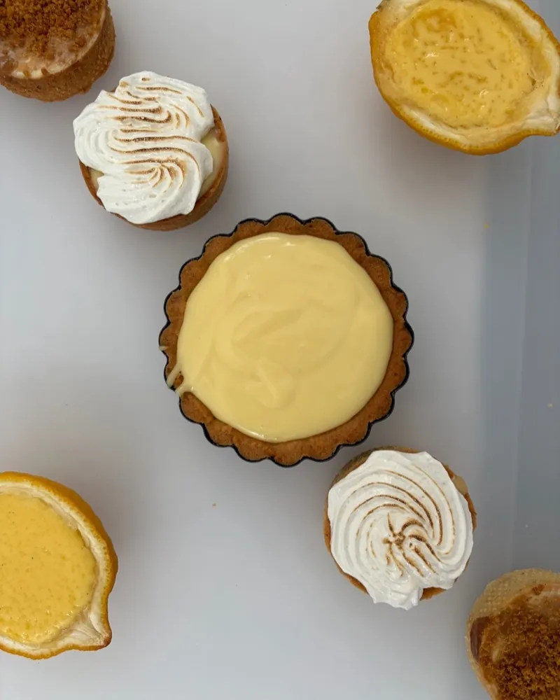 Seen from above: a lemon tart in the middle, surrounded by tartlets with meringue and Biscoff crumble and two lemon halves filled with yellow cream.
