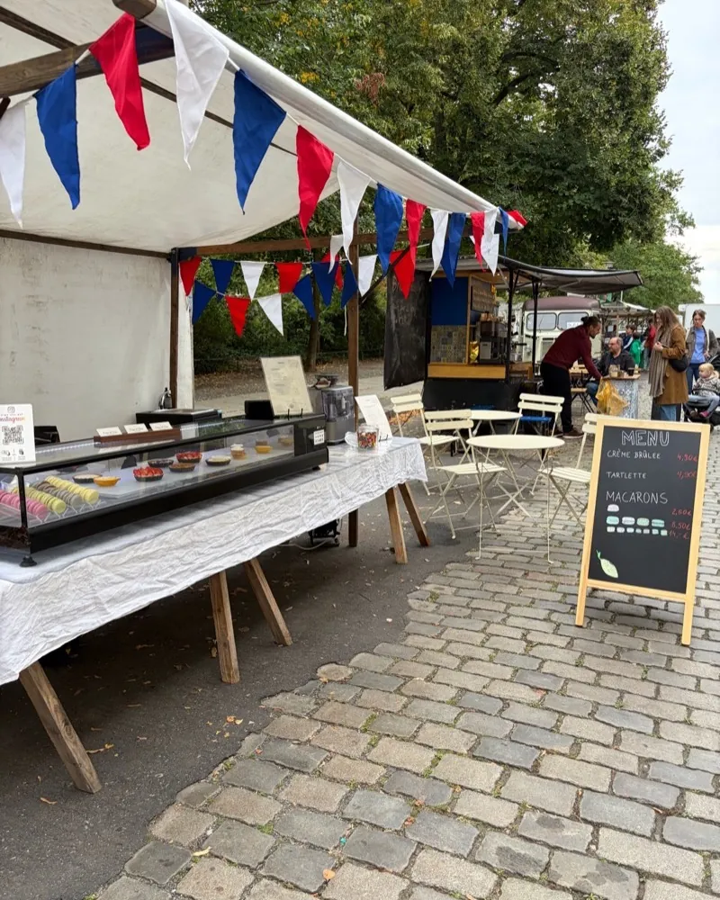 The Crème de Paris stall at the Bötzowmarkt with blue, white and red bunting, a display case and a menu board on the cobblestones.