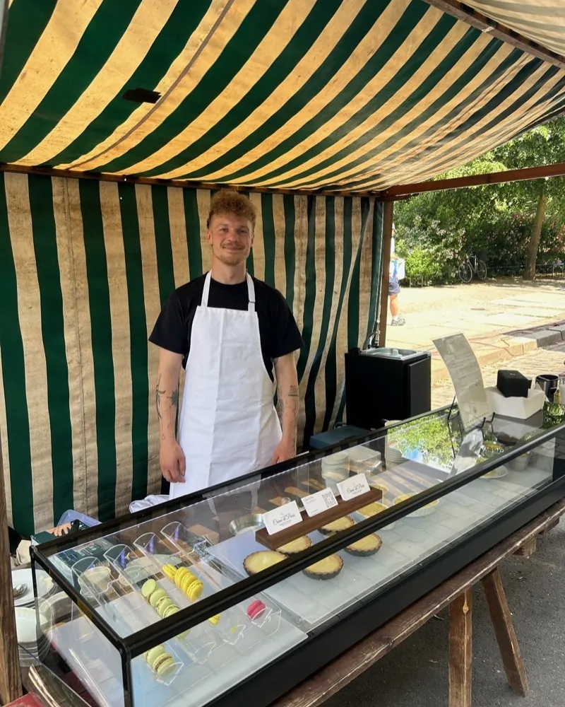 The Crème de Paris stall at the Bötzowmarkt: a man in a white apron behind a display case with macarons and tartlets, under a green-and-white striped awning.