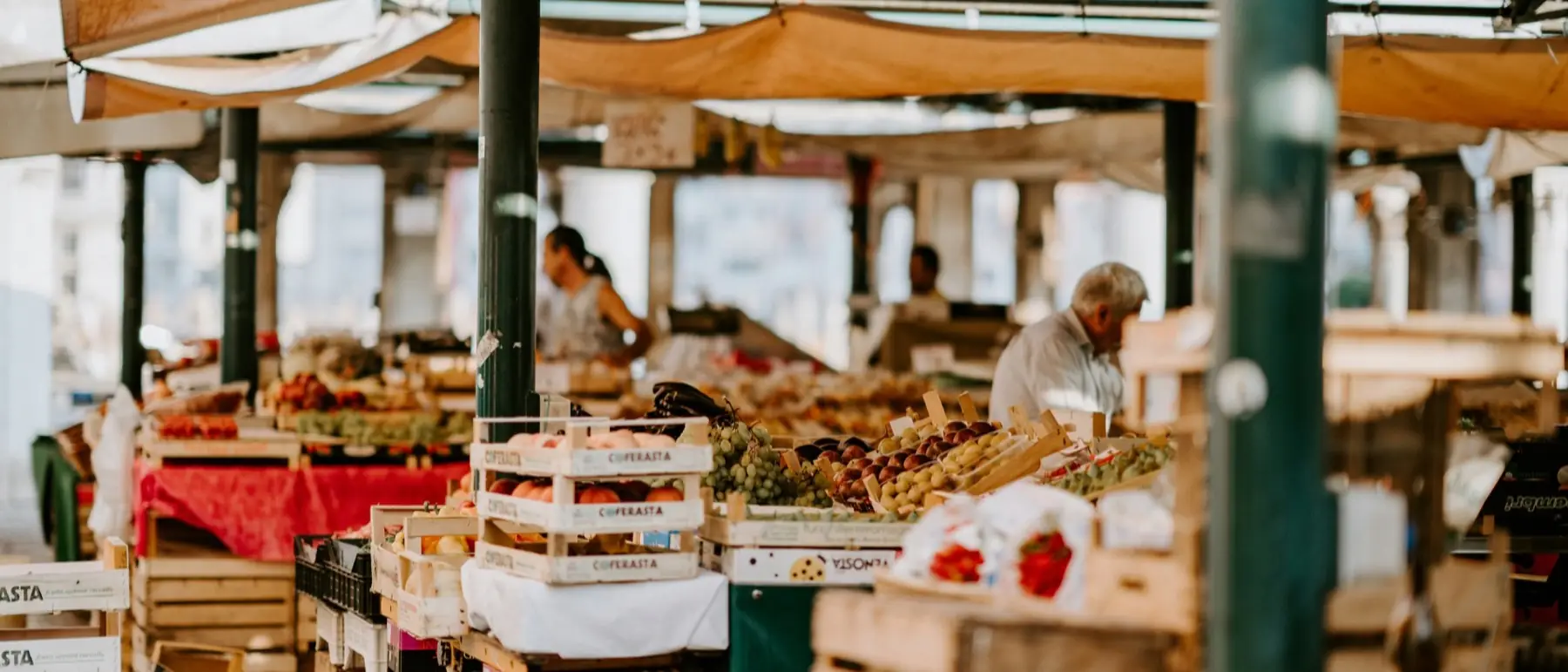 Lange Marktzeile unter Planen: Holzkisten mit Obst und Gemüse, im Hintergrund Händler an ihren Ständen.