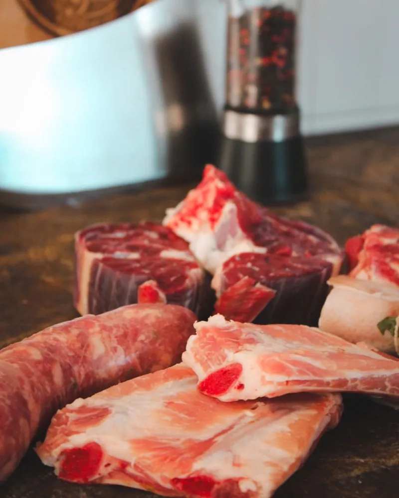 Raw meat and a sausage on a wooden board, a pepper mill behind them.