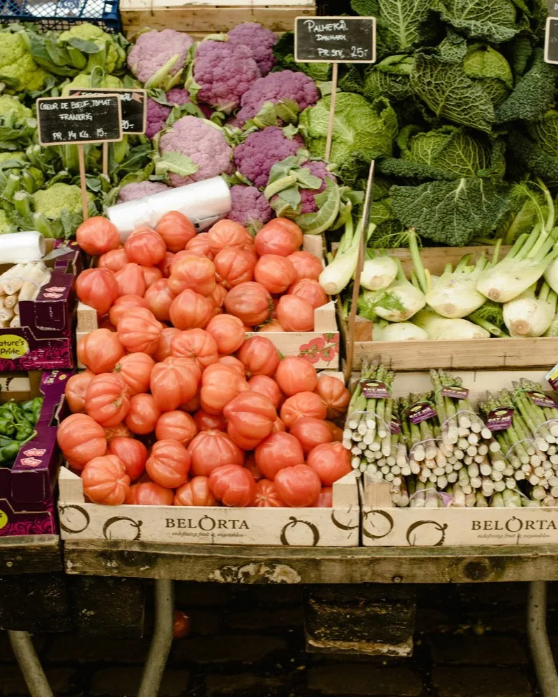 Market stall with wooden crates full of beef tomatoes, next to asparagus, fennel, cauliflower and savoy cabbage.