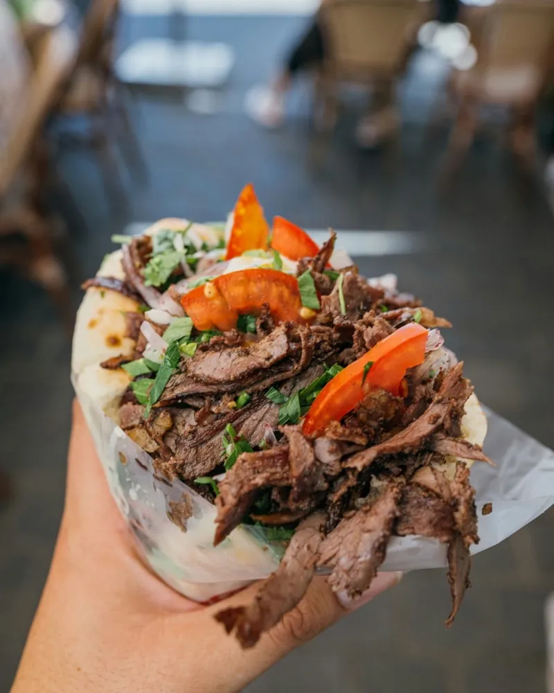 A hand holding a filled flatbread with meat, tomato and herbs.