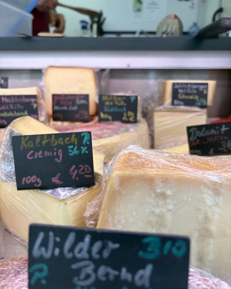 The Kochs Käsestandl cheese counter at the Bötzowmarkt with large wheels and labelled price tags.