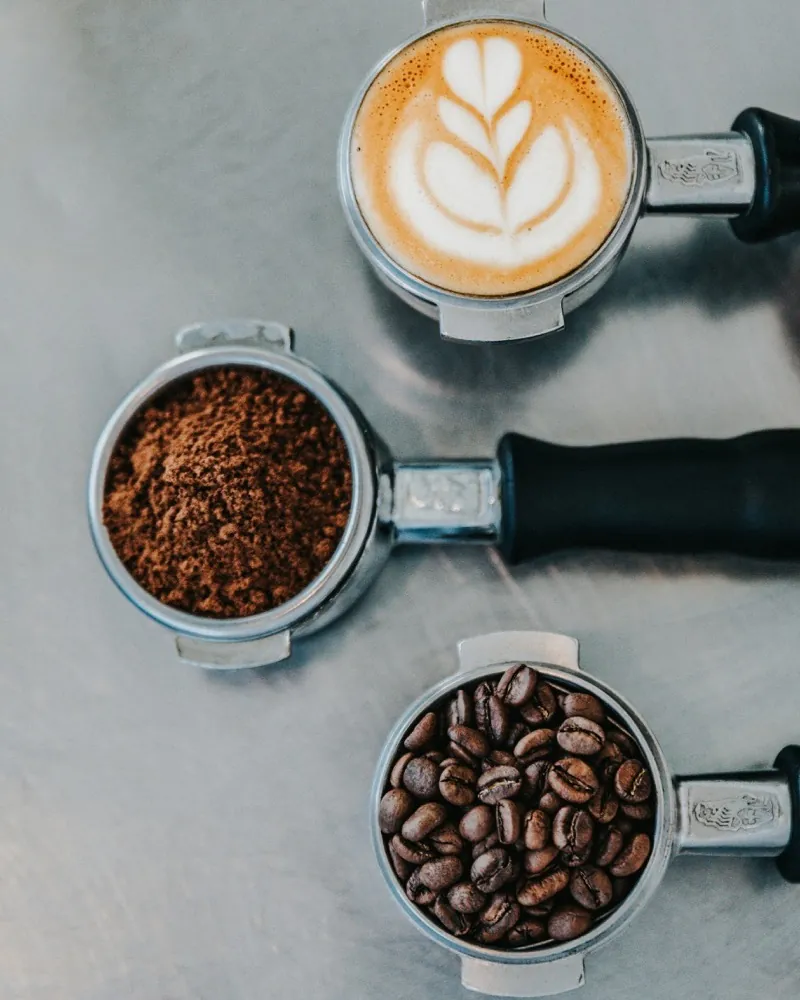 Three portafilters from above: a cappuccino with latte art, ground coffee and roasted beans.