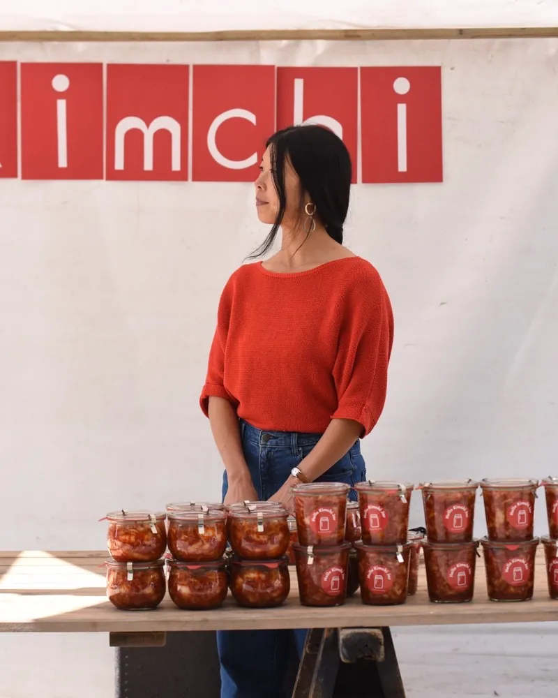 The owner of Meli's Homemade Kimchi at her stall at the Bötzowmarkt, jars of red kimchi stacked in front, a banner reading “kimchi” behind her.