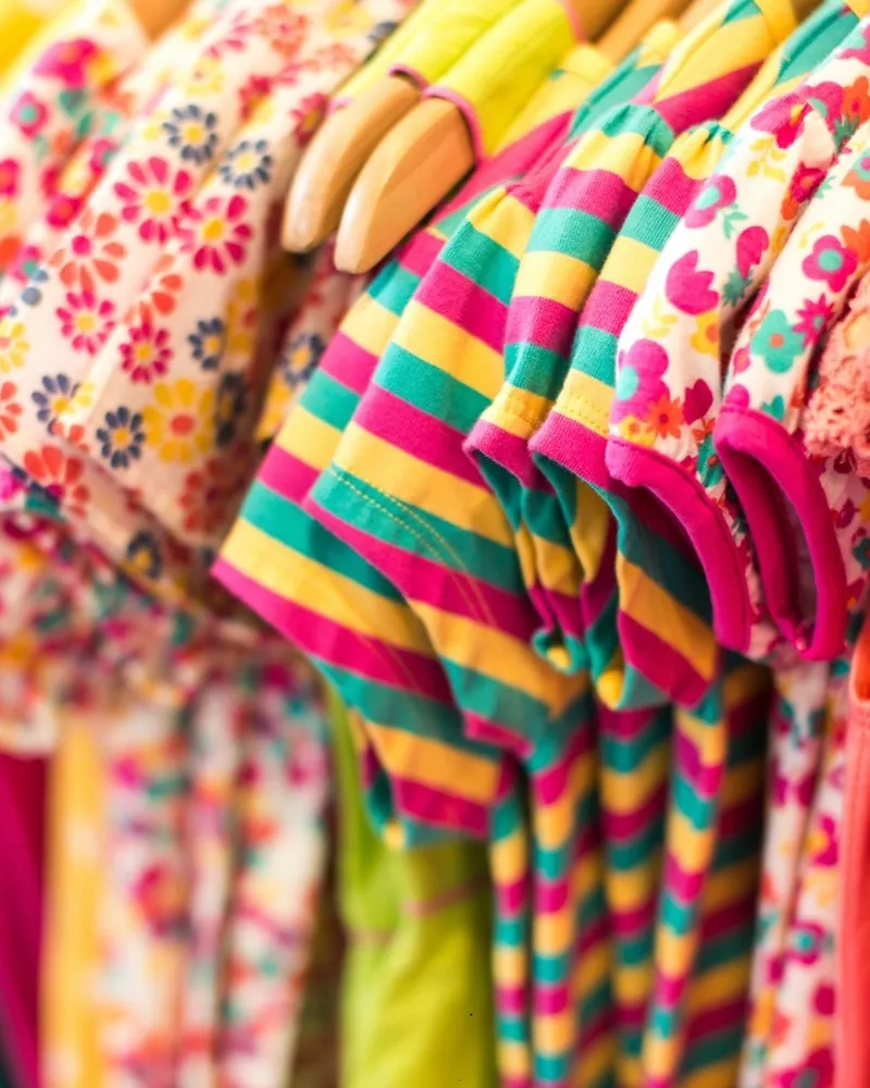 Colourful children's clothes packed close on wooden hangers: floral prints, striped shirts and frills.