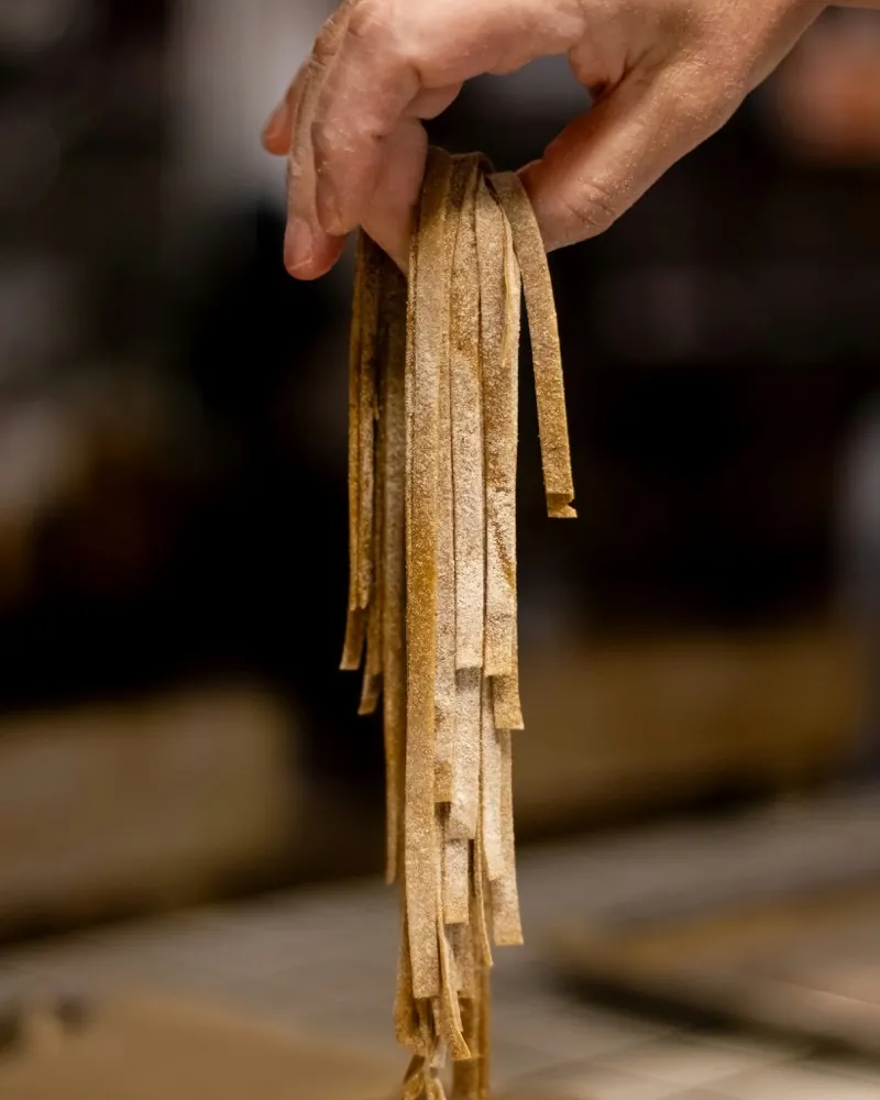 A hand holding up freshly cut ribbon pasta, the floured work surface below.