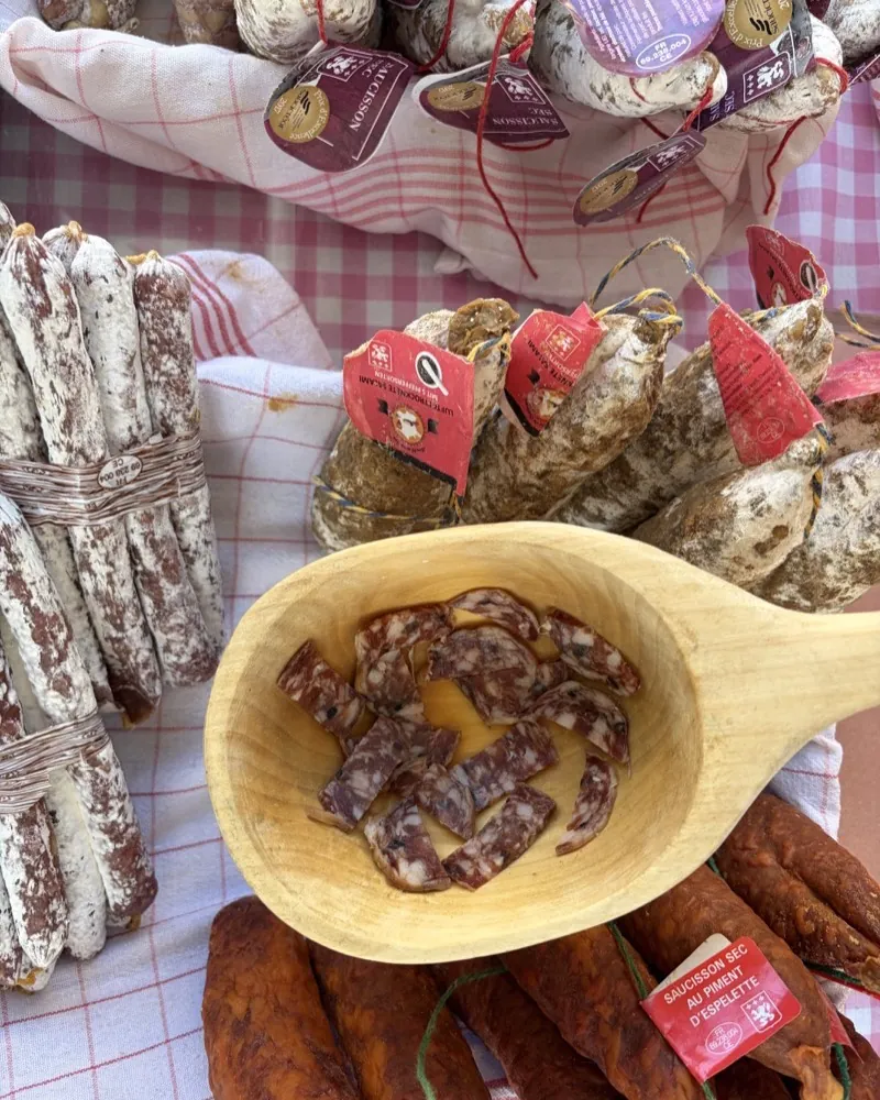 A wooden spoon with slices of French salami at the Marché de Catherine stall at the Bötzowmarkt, whole saucissons in a basket behind.
